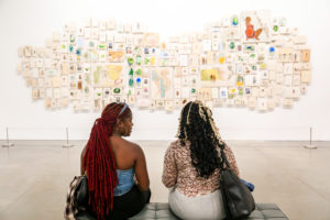 Two women sit on a bench in an art gallery, facing a large wall display of various small artworks and illustrations arranged in an organic pattern. The gallery has a modern, minimalist feel.