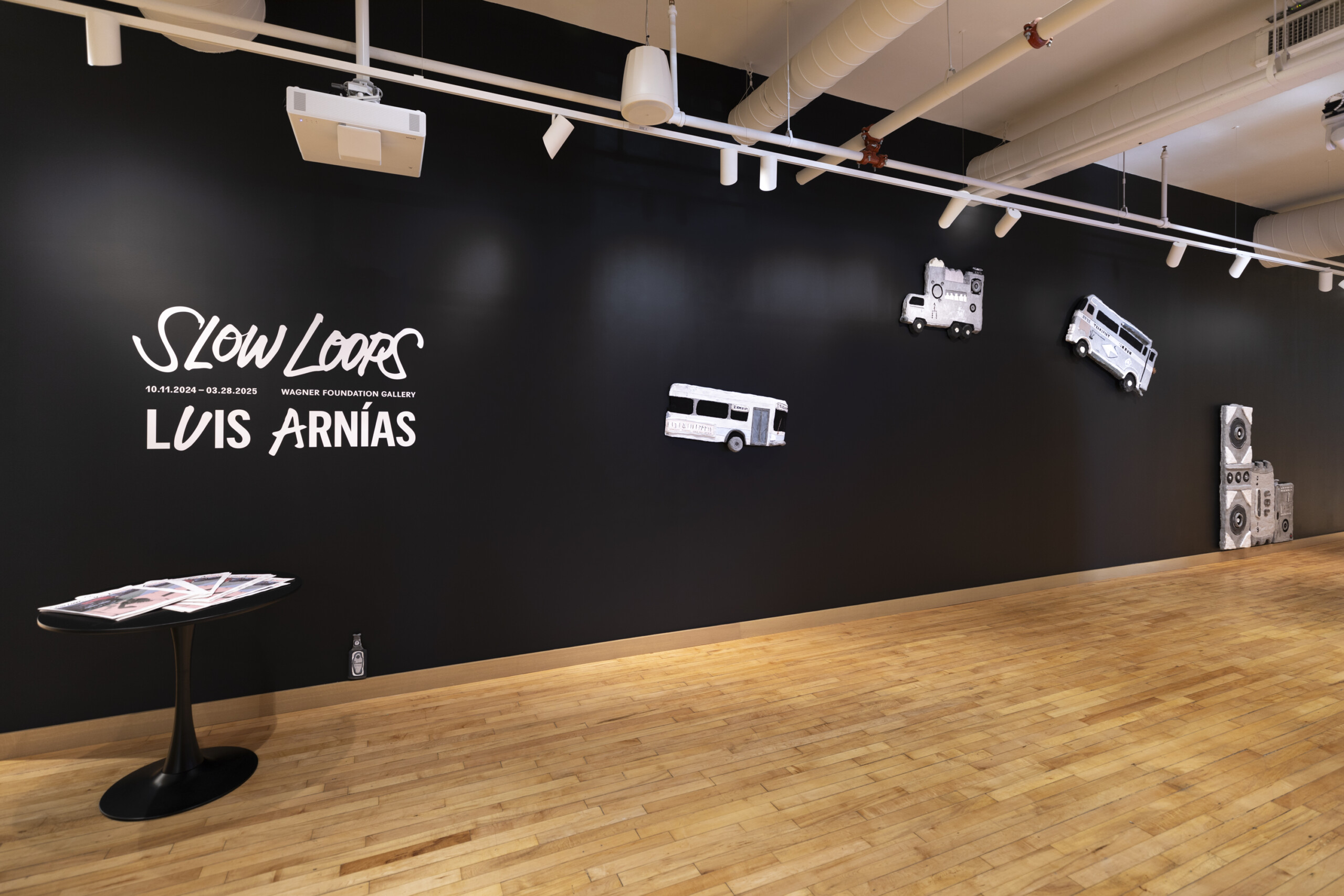 Gallery wall featuring the title Slow Loops, Luis Arnías and dates, with three white bus cutouts and a stack of printed materials on a small table.