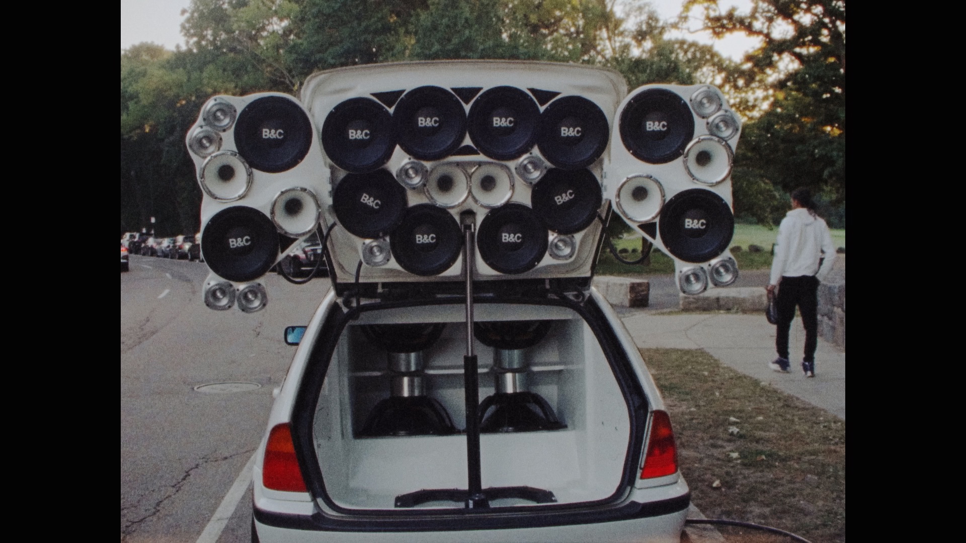 A small white car parked with its trunk open, revealing a large custom sound system with many speakers and subwoofers arranged inside and on the trunk lid. A person walks by in the background. Trees and a road are visible.