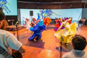 Dancers in brightly colored flowing dresses perform energetically on a stage with a brick floor, while musicians play in the foreground and an audience watches in the background.
