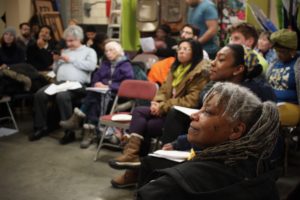 A diverse group of people sit in chairs, attentively listening and watching a presentation or event in an indoor community space. Some hold papers, and the atmosphere appears engaged and focused.