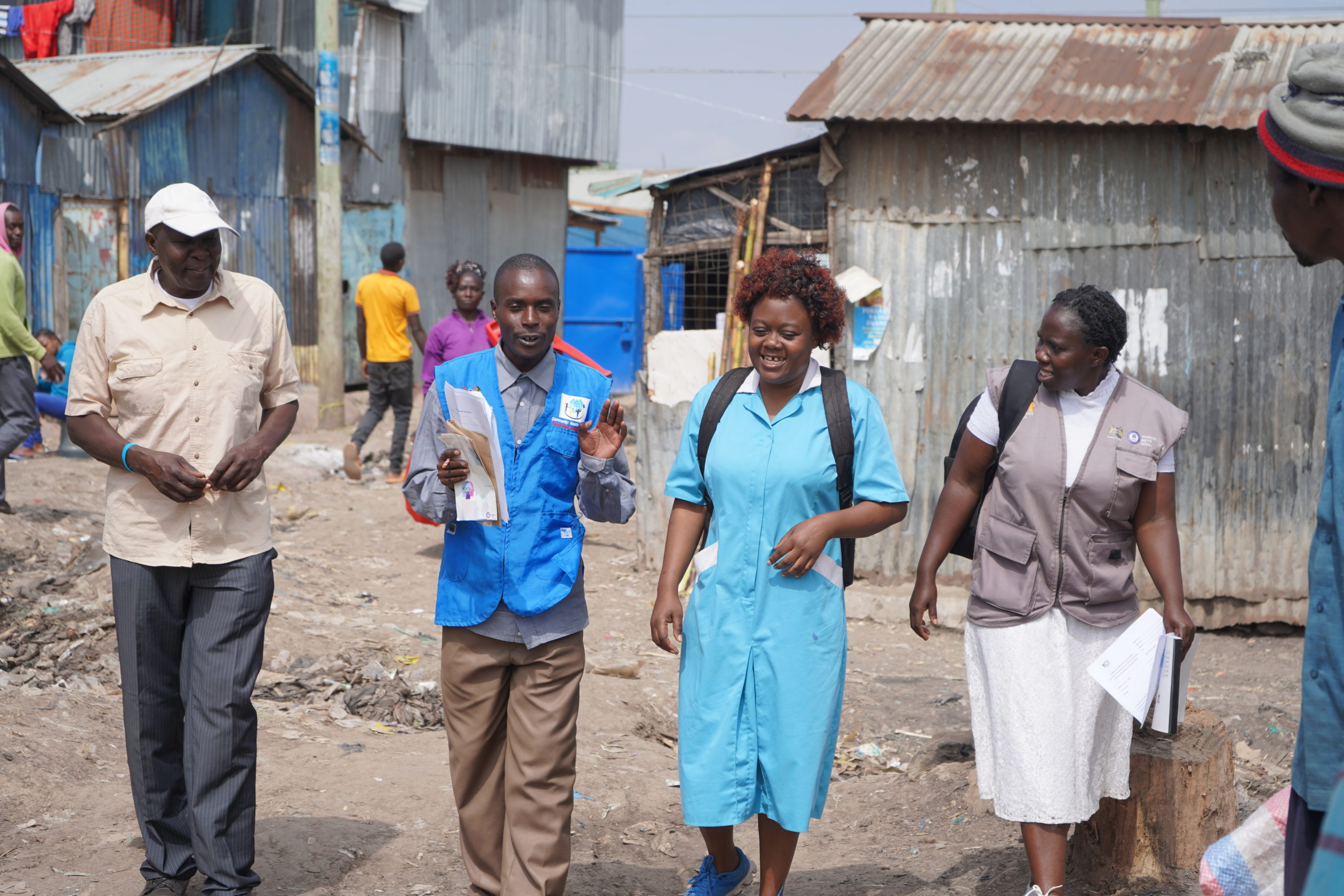 Four people walk and talk together through a village with corrugated metal buildings. Two wear uniforms and badges, carrying papers, appearing to be health or community workers. Others are visible in the background.