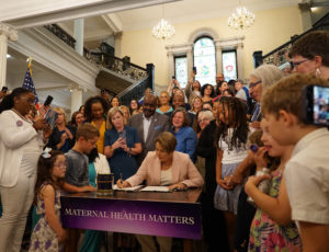 A diverse group gathers around a table with a “Maternal Health Matters” banner as a woman sits and signs a document, surrounded by children and adults in a grand, well-lit building.