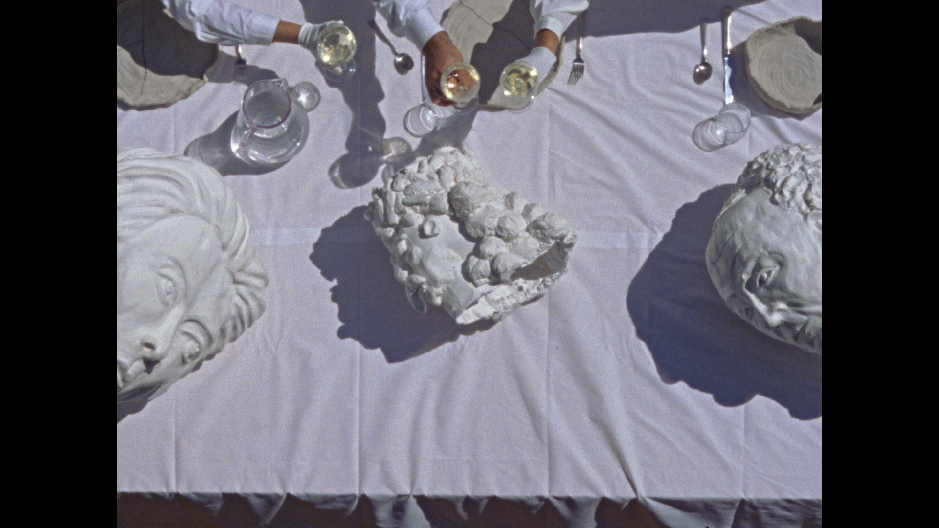 A white tablecloth holds three large sculpted heads, glassware, and plates. Two people in white sleeves pour clear drinks into glasses from above. Silverware is neatly arranged beside the plates.