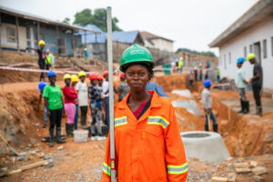 A woman in an orange safety uniform and green helmet stands at a construction site, holding a pole. Other workers wearing hard hats are visible in the background, working and talking near trenches and buildings.