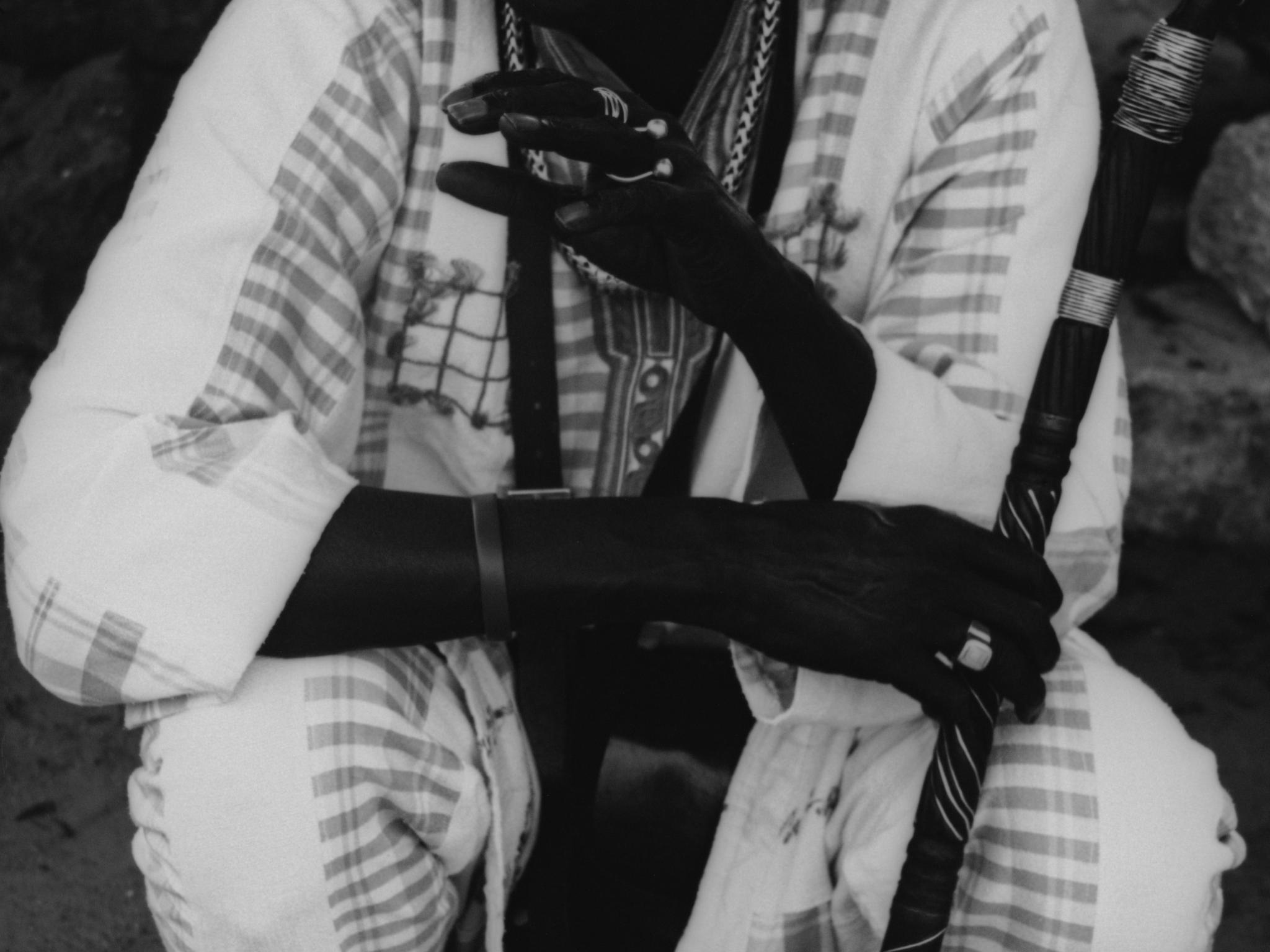 Black and white photo showing a person squatting, dressed in a patterned garment, holding a staff. The persons hands, adorned with rings and a bracelet, are prominently featured in the foreground. Face and surroundings are partially obscured.