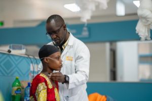 A doctor in a white coat uses a stethoscope to examine a patient wearing a patterned dress and nasal cannula in a medical clinic.