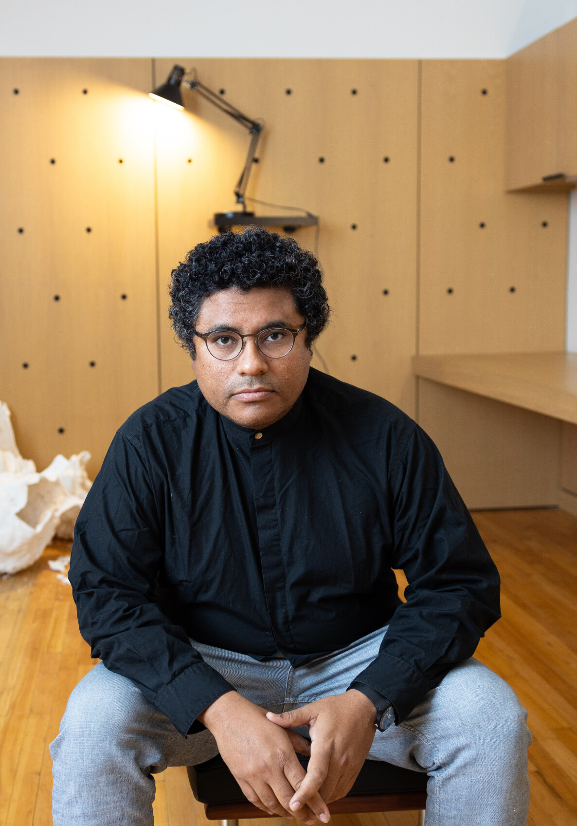 A person with curly hair and glasses, wearing a black shirt and light gray pants, sits on a chair in a modern office with wood-paneled walls, a lamp, and a desk in the background.