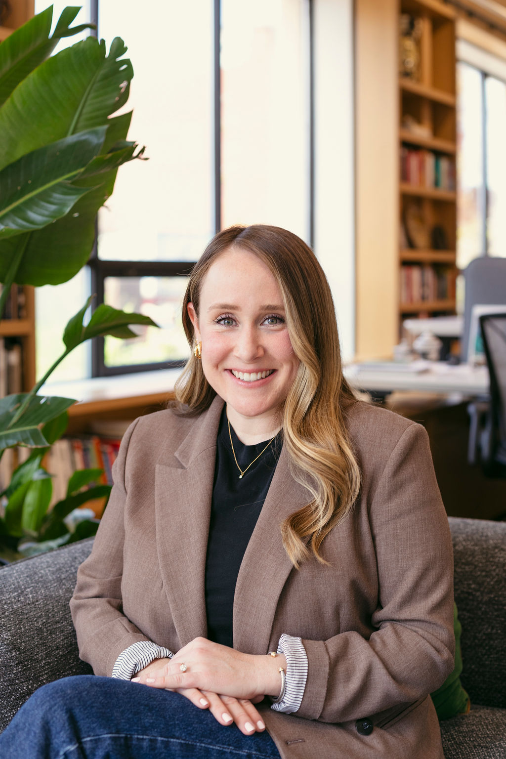 A woman with long blonde hair, wearing a brown blazer and black top, sits smiling on a gray chair in a bright office with large windows, green plants, and bookshelves in the background.