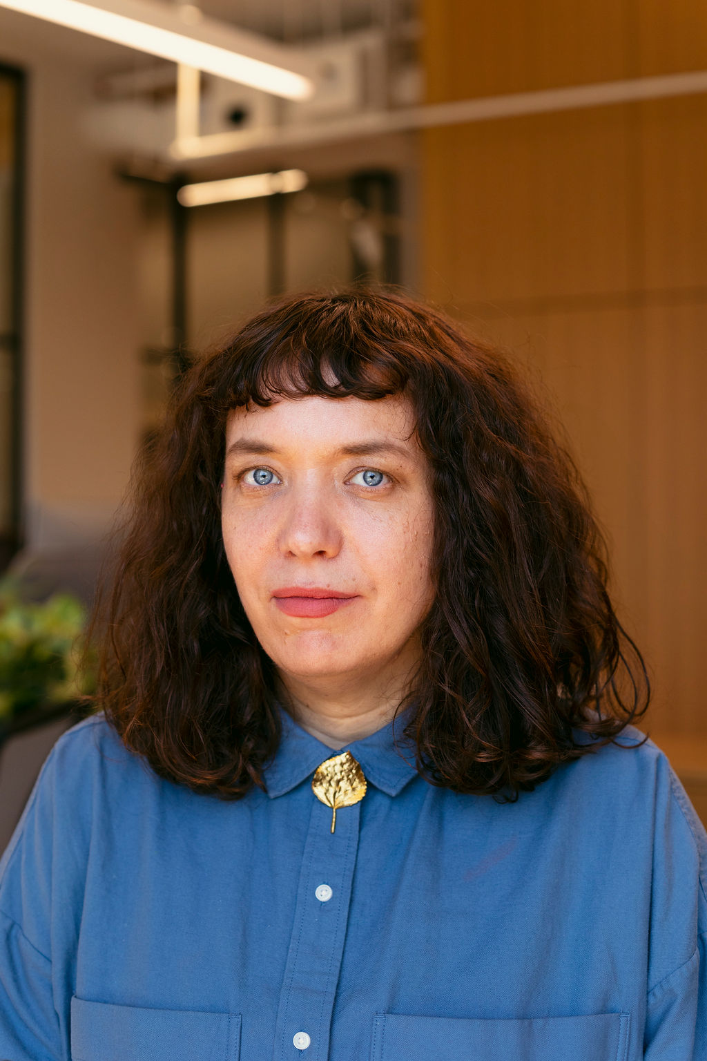 A person with wavy brown hair and blue eyes, wearing a blue button-up shirt with a gold brooch at the collar, looks at the camera in a modern indoor setting with warm lighting and blurred background.