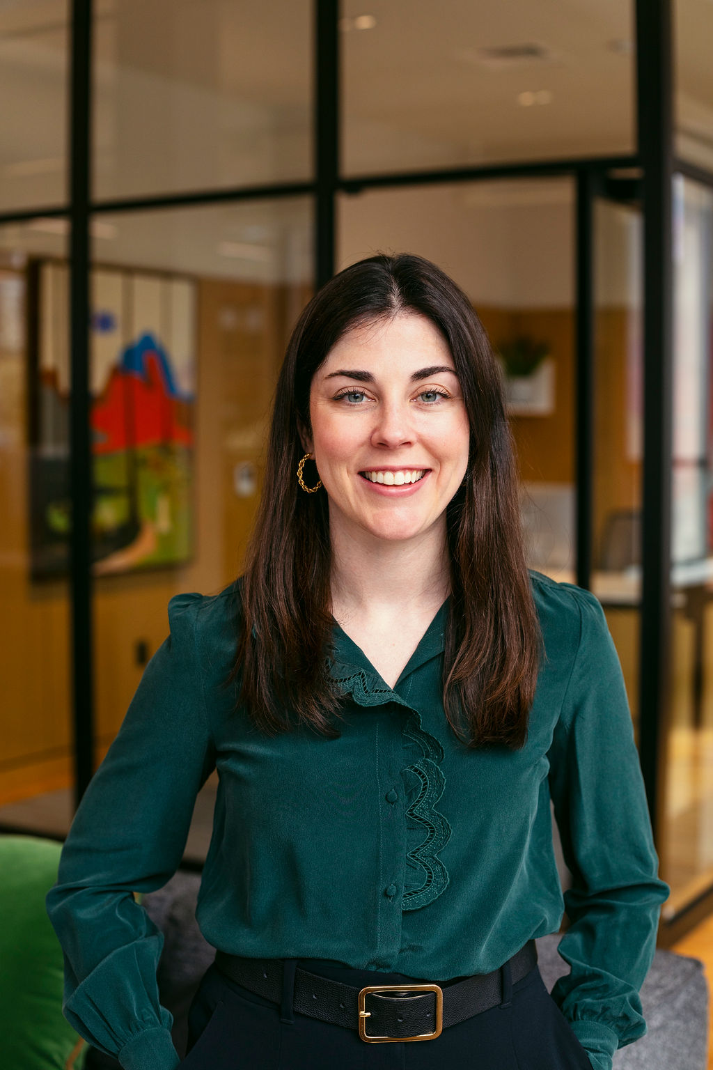 A woman with long brown hair, wearing a dark green blouse and black pants with a belt, stands smiling in a modern office with glass walls and colorful art in the background.