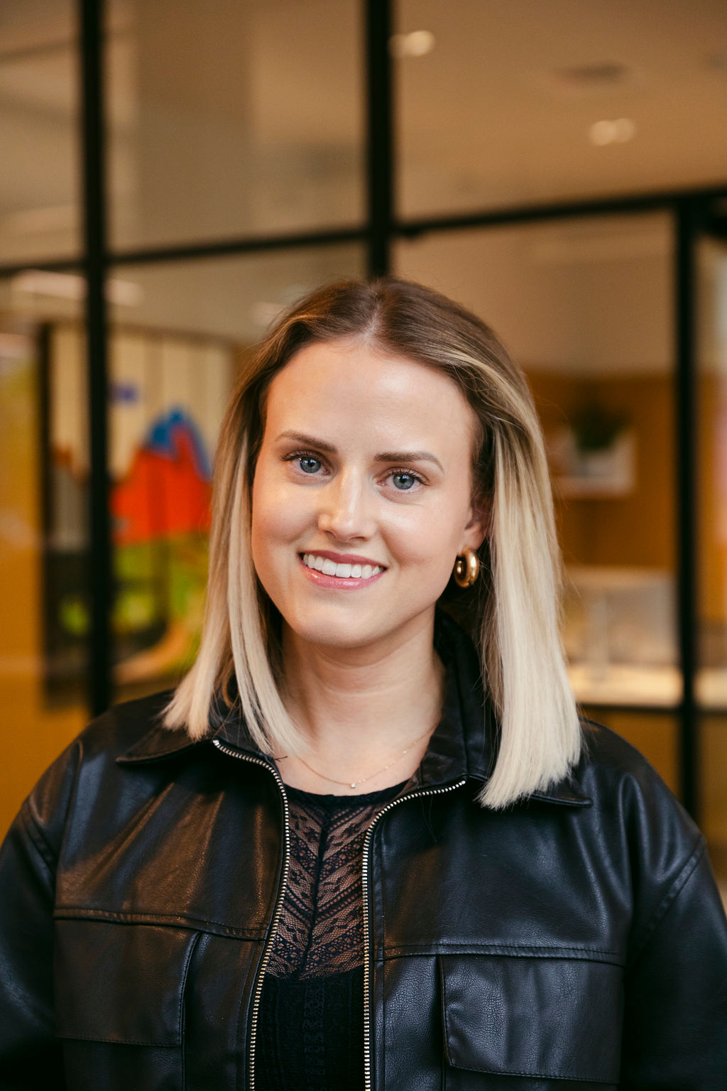 A woman with straight blonde hair, wearing a black leather jacket and a black lace top, smiles at the camera. She is standing indoors in a modern office space with glass walls and blurred background details.