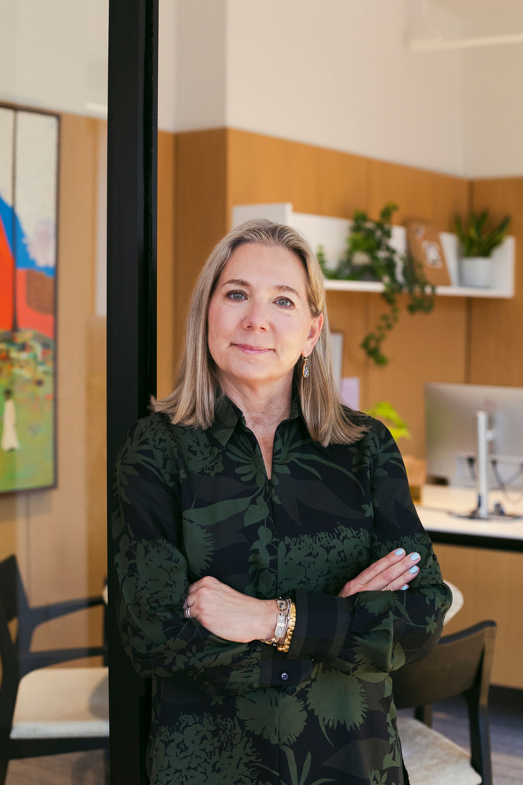A confident woman with long blonde hair, wearing a dark patterned blouse, stands with arms crossed in a modern office with wooden walls, plants, and a computer on the desk behind her.