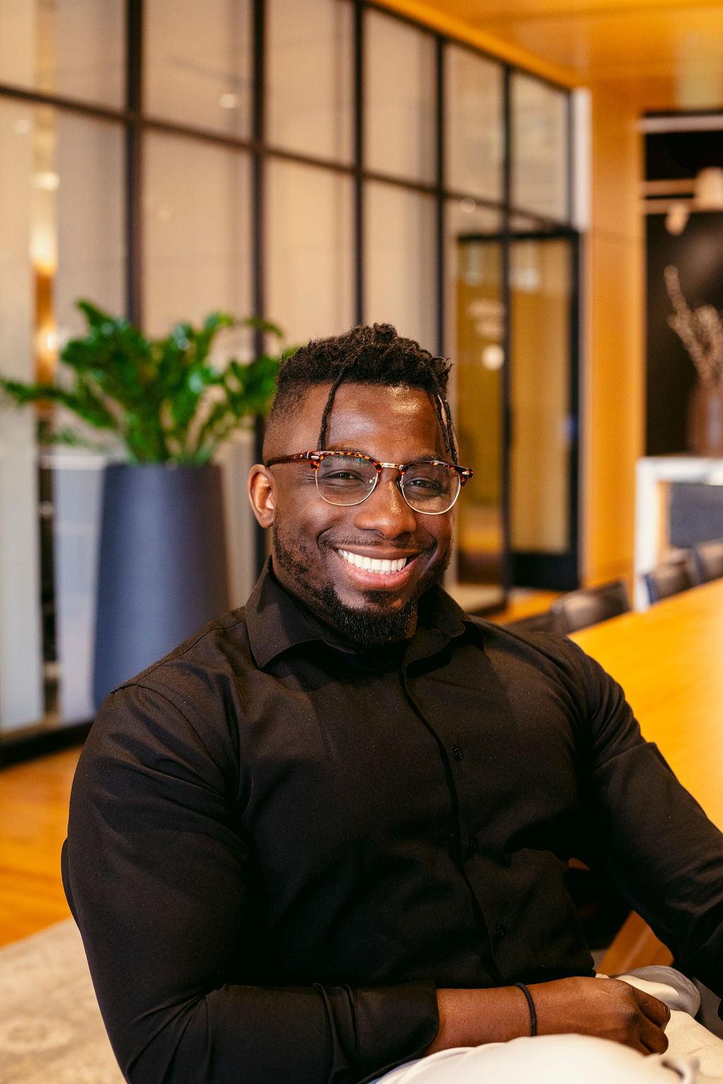 A smiling man wearing glasses and a black shirt sits at a wooden table in a modern office with large windows and plants in the background.