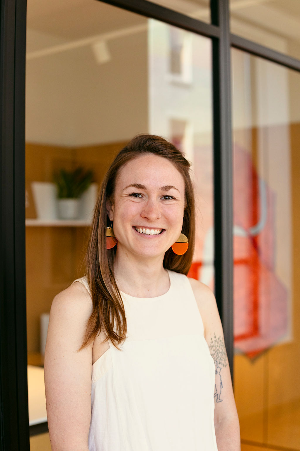 A woman with long brown hair, wearing a sleeveless white top and orange earrings, smiles while standing indoors near a glass wall. A tattoo is visible on her upper arm, and there are shelves and plants in the background.