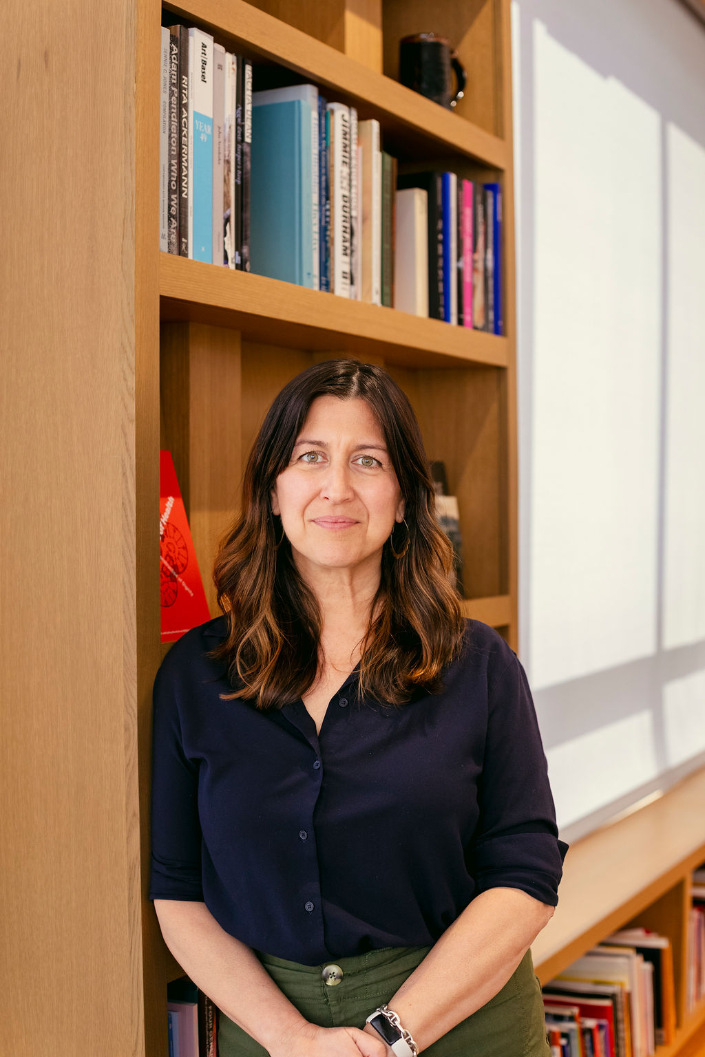 A woman with wavy brown hair wearing a navy blouse stands in front of a wooden bookshelf filled with books, with sunlight streaming through a window beside her.