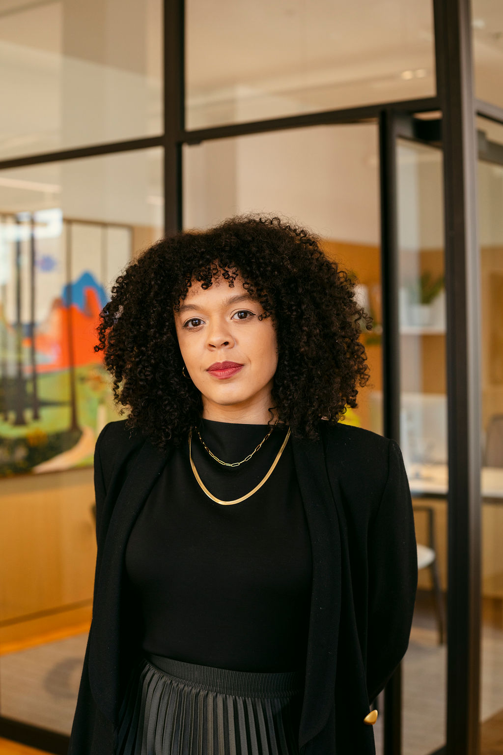 A woman with curly hair, wearing a black top, black blazer, and gold necklace, stands in a modern office with glass walls and contemporary décor.