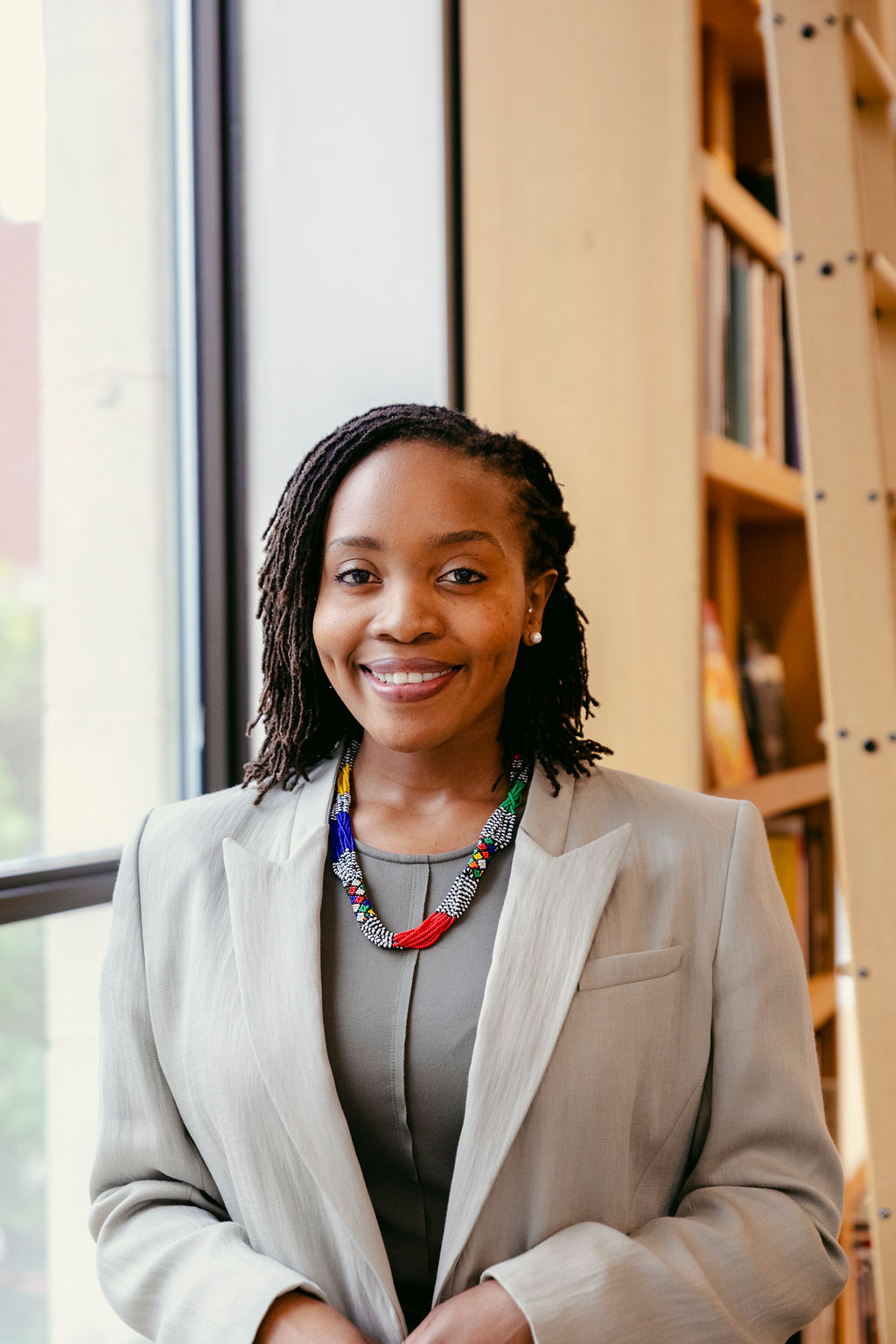 A woman with shoulder-length braided hair, wearing a light gray suit and a colorful beaded necklace, stands smiling in front of a window and bookshelves.