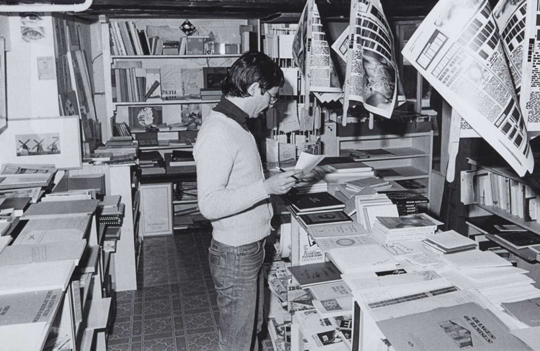 A person wearing glasses and a sweater stands in a small bookstore or library, browsing through a book. The space is filled with shelves and tables stacked with books and papers, with posters hanging overhead.