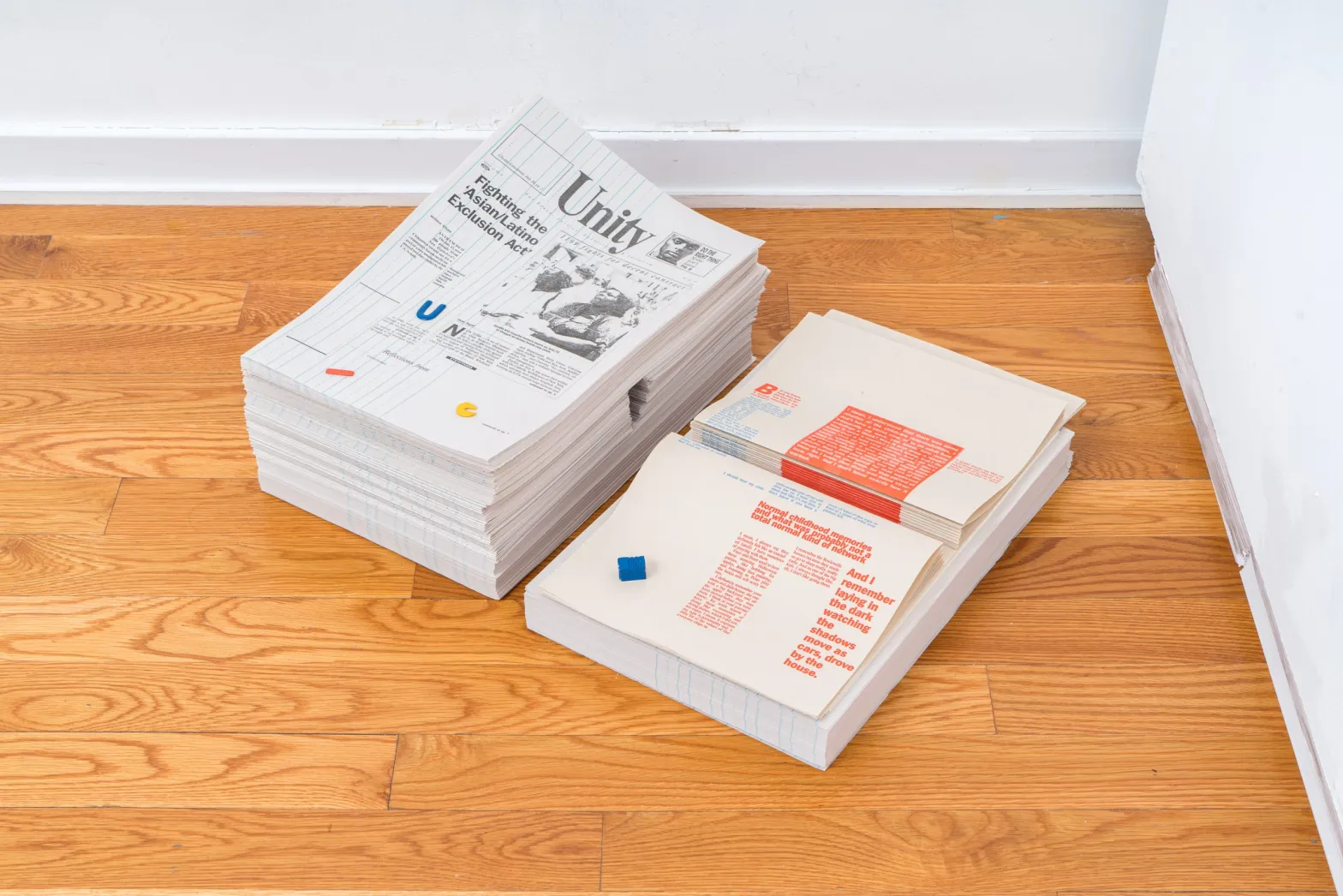 Two stacks of newspapers or printed materials rest on a wooden floor near a white wall. One stack has a visible front page titled Unity with a black-and-white photo; the other shows pages with red and blue text blocks.