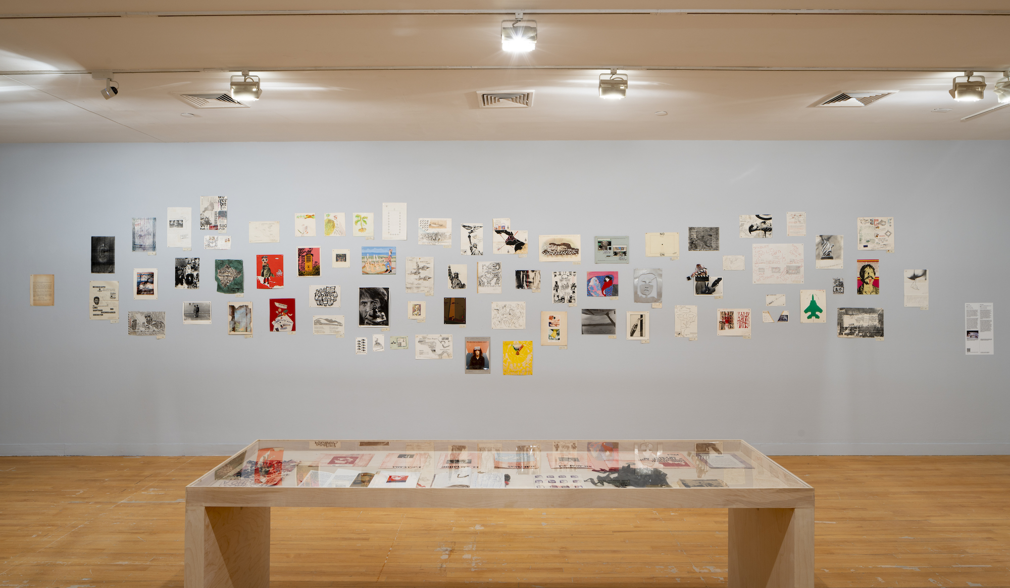 A gallery exhibit featuring a wall with various artworks, notes, and photographs arranged in a collage, and a display table beneath holding more documents and images, all on a light blue wall and wooden floor.