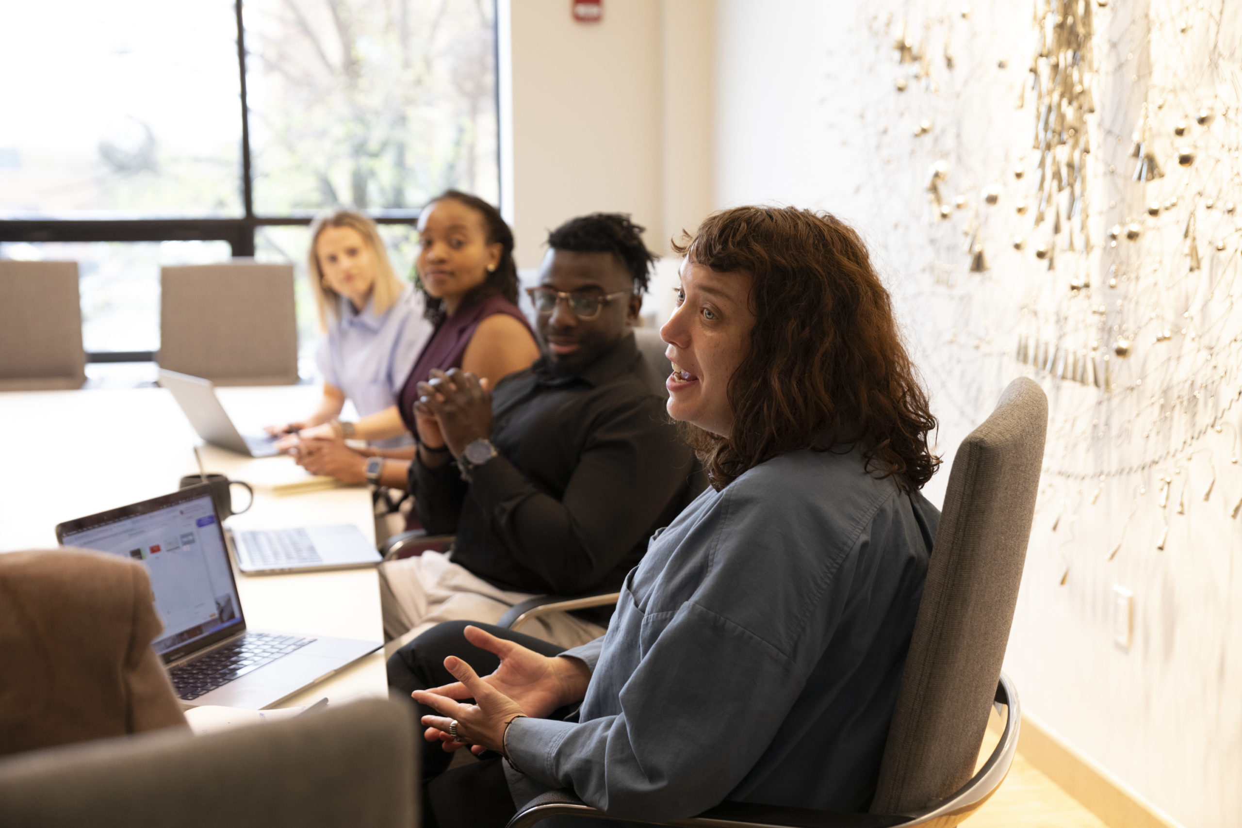Four people sit at a conference table with laptops, engaged in a discussion. One person in front, wearing a blue shirt, gestures while speaking. The setting is a bright, modern office with natural light.