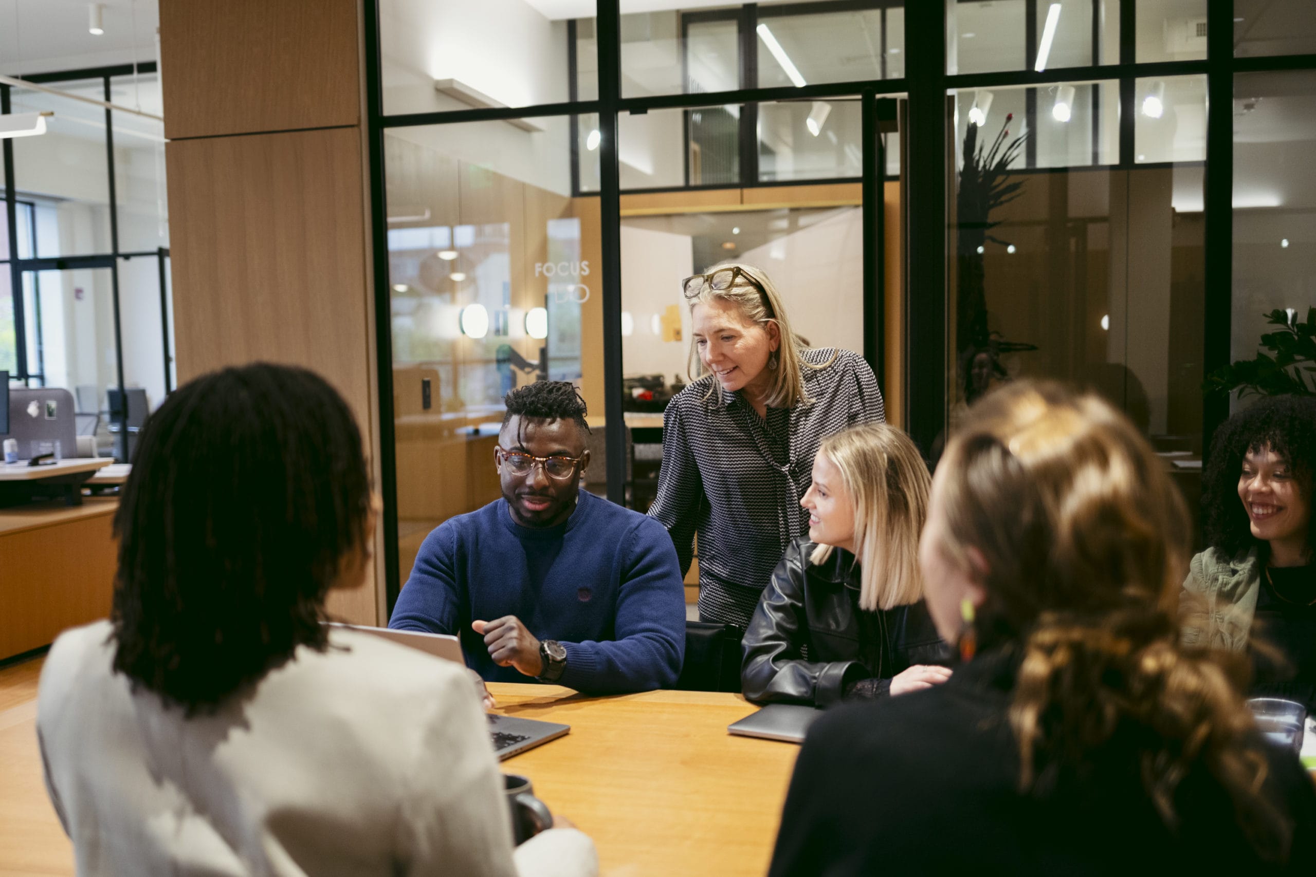 A group of five people sits around a table in a modern office, having a discussion. One person stands and leans over, engaging with the group, while the others listen and smile. Laptops and notebooks are on the table.