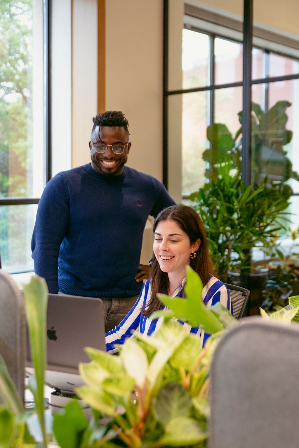 Two colleagues work together in a modern office with large windows and lots of plants; a smiling man stands beside a woman who is sitting and working on a laptop, both appearing engaged and happy.
