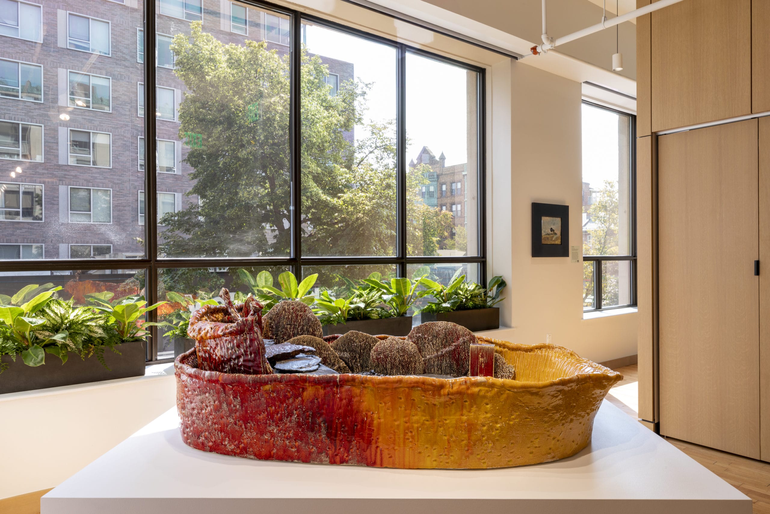 A large, colorful ceramic sculpture resembling a boat sits on a white pedestal in a bright room with large windows, potted plants, and wooden panels. An urban building and trees are visible outside.