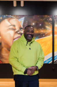 A man wearing a bright green hoodie smiles while standing indoors with his hands clasped. Behind him is a colorful mural featuring a joyful child and vibrant, cosmic-themed art.