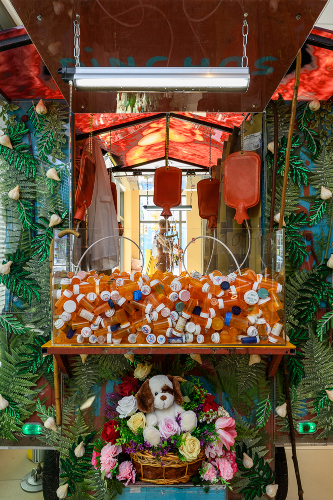 A claw machine filled with orange prescription pill bottles, surrounded by fake ferns and leaves. At the bottom sits a stuffed dog toy in a basket with pink flowers. In the background, a skeleton is visible.