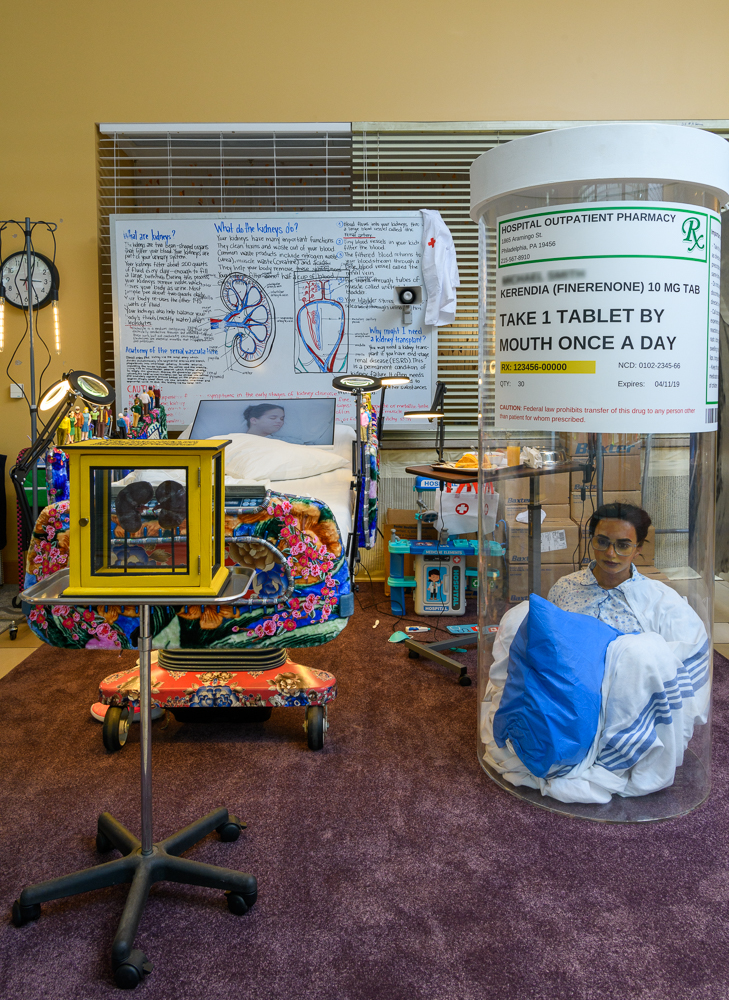 A room displays a hospital bed with medical equipment, a mannequin in a large pill bottle labeled for daily medication, a framed picture on a stand, and detailed diagrams on the wall explaining kidney function.