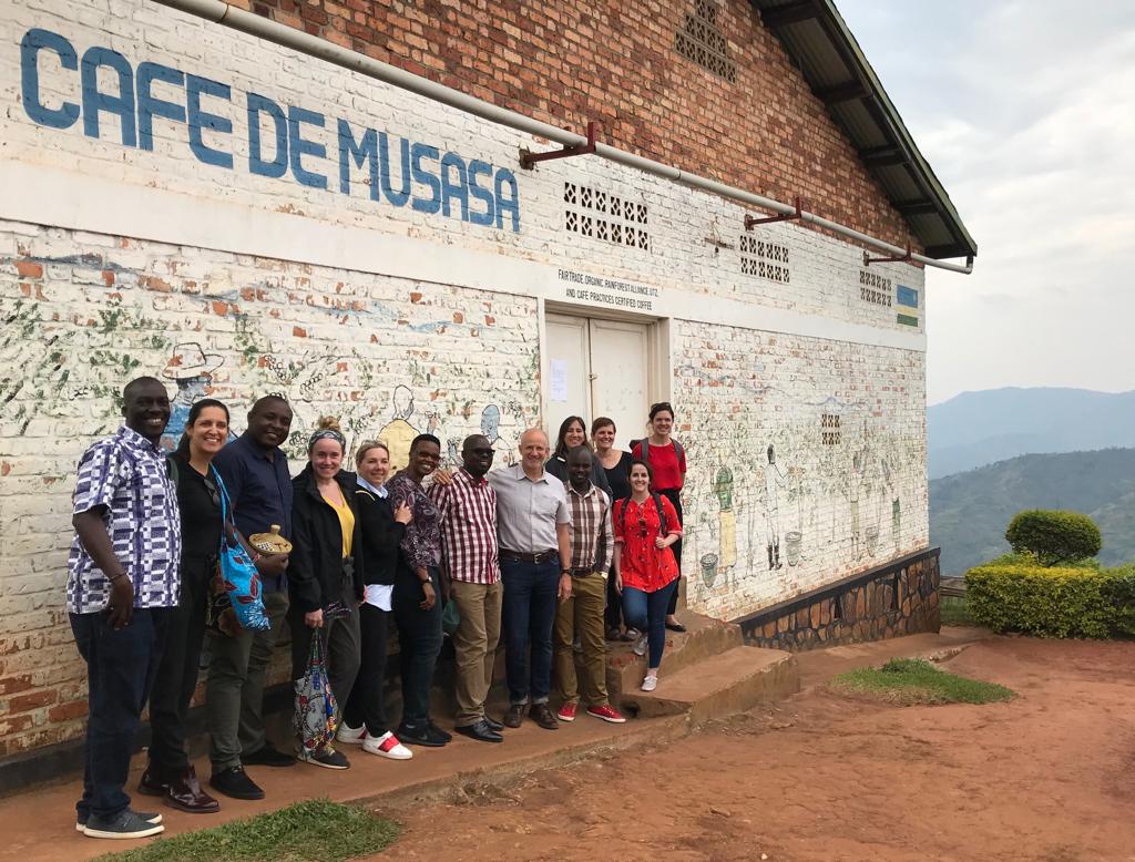 A diverse group of people stands smiling in front of a brick building with Café de Musasa painted on the wall. The background features a scenic, hilly landscape and greenery.