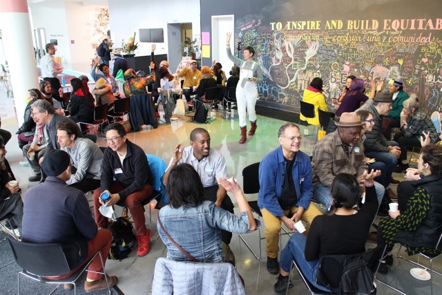 A diverse group of people sitting in small circles, talking and smiling, in a bright room with a colorful chalkboard wall behind them that reads TO INSPIRE AND BUILD EQUITABLE and features various drawings and words.