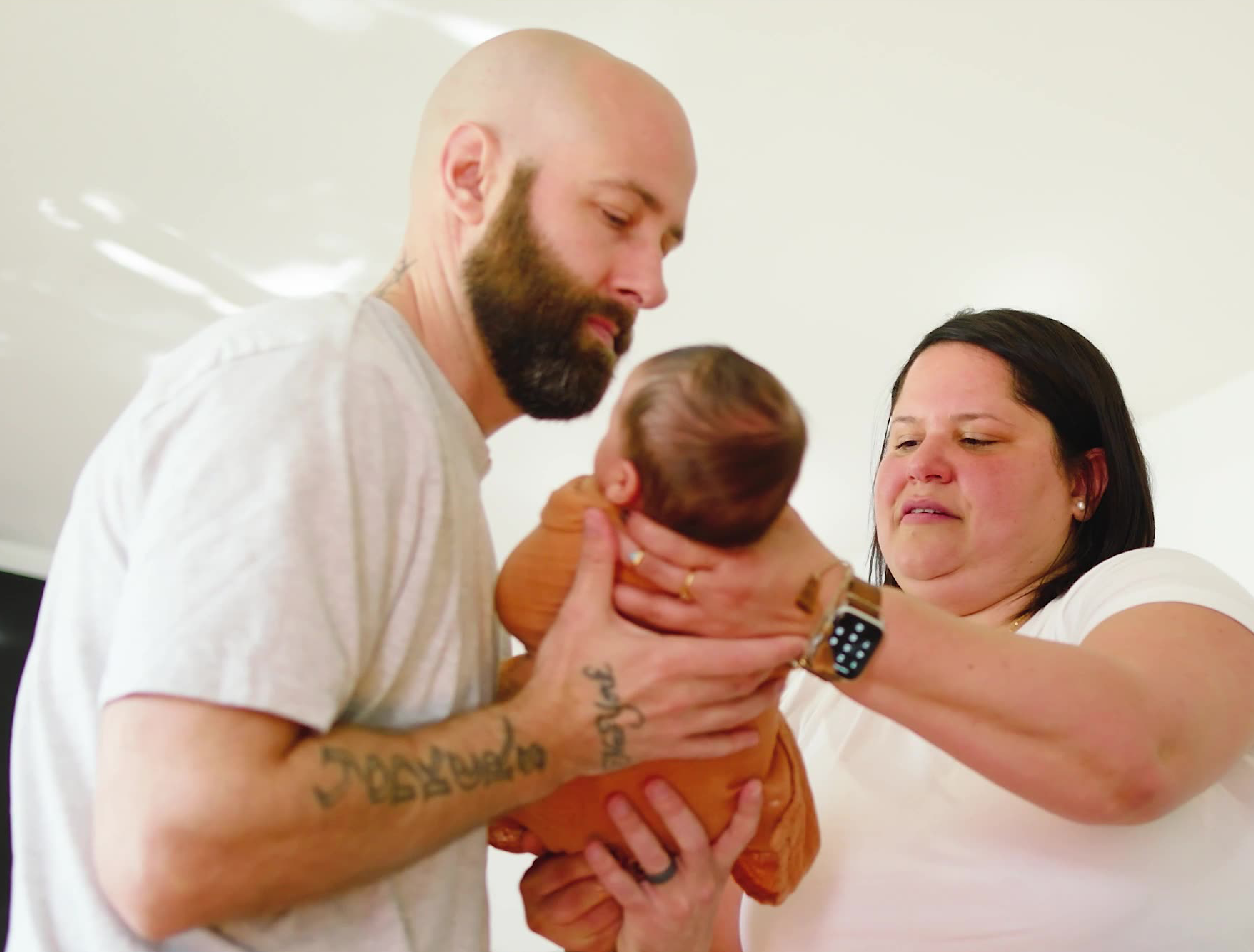 A bearded man and a woman gently hold a baby together. The man looks at the baby with care while the woman supports the baby and smiles softly. Both adults appear attentive and loving.