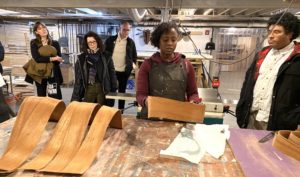 A woman wearing an apron demonstrates curved wooden pieces to a group of attentive adults inside a workshop, with tools and materials visible on the worktable and shelves in the background.
