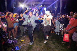 Two men dance energetically in the center of a lively outdoor crowd at night, surrounded by people cheering, filming with phones, and smiling under colorful stage lights. A band performs on a lit stage in the background.