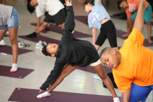Children are practicing yoga indoors on mats, stretching with arms up and legs apart. One child in a black sweatshirt is in the center, smiling, while others around also hold the pose. Shoes are off to the side.