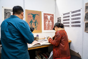 Two people view art books at a small desk in a gallery or studio. Artworks and black-and-white photo strips are displayed on the surrounding white walls. Shelves with more books are visible under the desk.