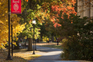 A tree-lined campus path at Bard College in autumn, with colorful foliage, benches, a lamppost, and a red Bard banner. People are walking and sitting, enjoying the sunny fall day.