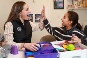 A woman and a young girl smile and give each other a high five while sitting at a table with colorful sensory toys and playdough. They appear happy and engaged in playful activity together.