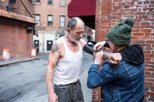 A man in a white tank top and gray pants receives an injection in his arm from another person wearing a green knit hat and denim jacket in an urban alleyway with brick buildings.