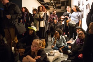 A diverse group of people, some seated on the floor and others standing, attentively watch something off-camera in an indoor art gallery setting with artwork displayed on the walls.