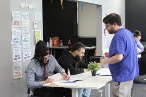 Two men sit at a table writing, while a man in a blue shirt stands and speaks to them. Papers, pencils, and a small potted plant are on the table. Posters reading “Said No Boss Ever” hang on the wall nearby.
