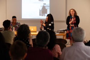 A woman stands and smiles holding a microphone while two people sit at a table; one claps and the other smiles. An audience watches and a projected image is displayed on the wall behind them.