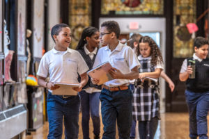 A group of elementary school children walk and talk in a hallway, carrying notebooks and papers. They are smiling and wearing school uniforms. The background shows classroom doors and colorful decorations.