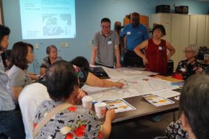 A group of adults gathers around a table covered with maps and documents, discussing and pointing at the papers. A presentation with charts is projected on the wall behind them.