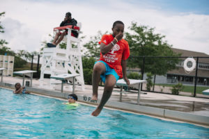 A boy in a red shirt and blue shorts holds his nose as he jumps into a swimming pool. A lifeguard watches from a tall chair. Another child is swimming in the water nearby. Trees and a building are in the background.