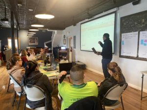 A man stands and presents information from a large screen to a group of seated adults in a classroom or meeting room. Attendees listen, take notes, and use laptops. A whiteboard with sticky notes is visible.