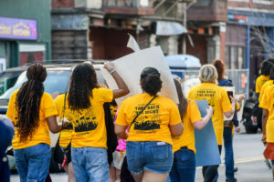 A group of people wearing matching yellow Take Back the Night t-shirts walk together down a street, some holding signs, participating in a community march.